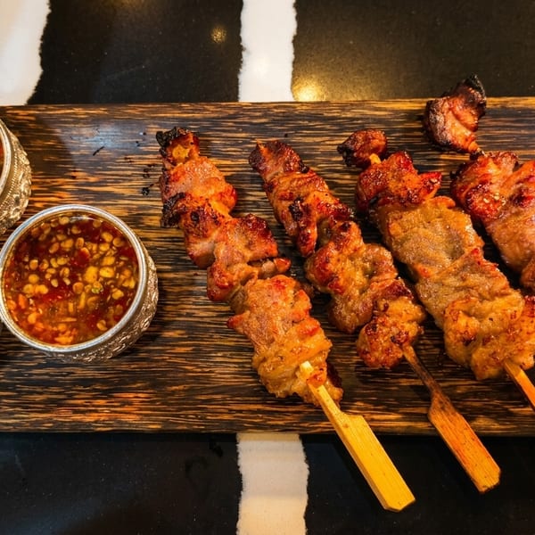 Grilled pork skewers with three dipping sauces on a wooden tray, against a black and white background.