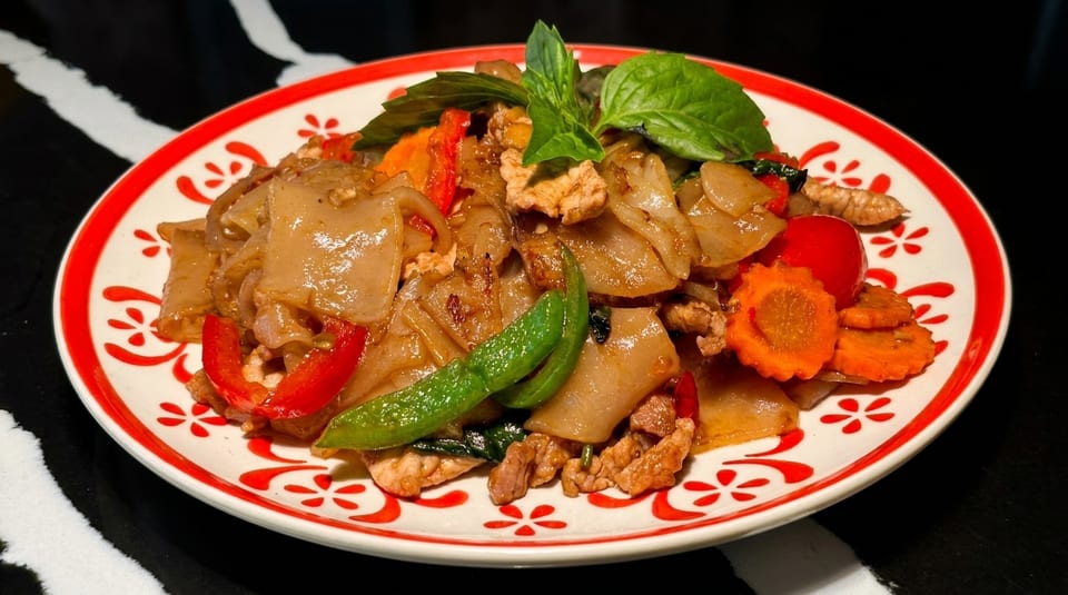 Pad See Ew with wide noodles, chicken, red peppers, carrots, and basil on a white plate.